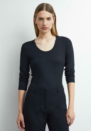 Woman with straight blond hair wearing a black long-sleeve scoop neck top and black pants standing against a white background.