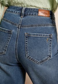 Blauwe denim jeans met een hoge taille, voorzien van koperen stiksel en een bruin leren label aan de achterkant. Twee achterzakken met kleine borduursels.