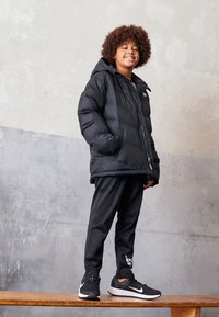 Chaqueta de plumas negra con capucha, combinada con pantalones deportivos negros y zapatillas negras con un logo blanco. De pie sobre una superficie de madera.