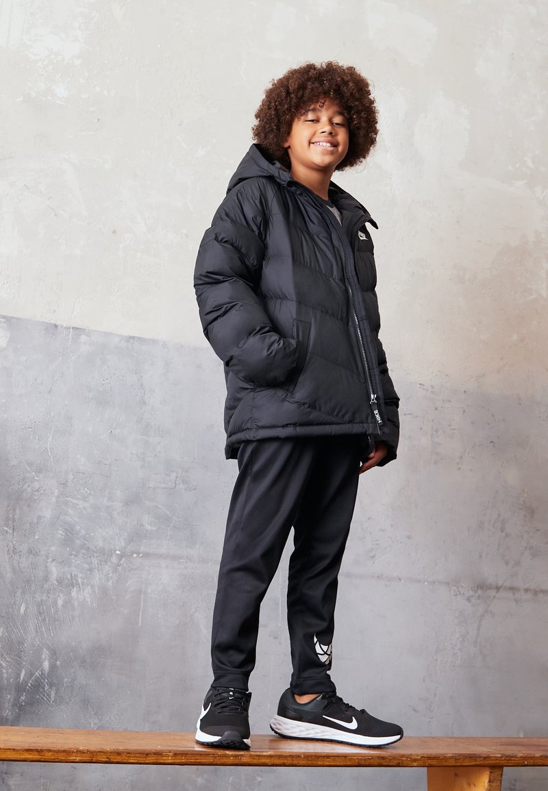 Chaqueta de plumas negra con capucha, combinada con pantalones deportivos negros y zapatillas negras con un logo blanco. De pie sobre una superficie de madera.