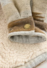 Zachte gebreide handschoenen met bruine en beige kleurvlakken en een klein rond metalen label waarop "Next Originals Since Eighty Two" staat, liggen op fleece stof.