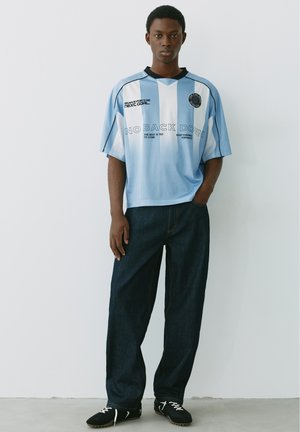 Giovane uomo in piedi con una mano in tasca, indossa una camicia oversize a righe azzurro chiaro e bianco con scritta e jeans scuri su sfondo semplice.