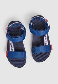 Modré sandály s nastavitelným páskem, červenými akcenty a bílými pásky s logem na černých strukturovaných stélkách, zobrazené z vrchu na bílém pozadí.