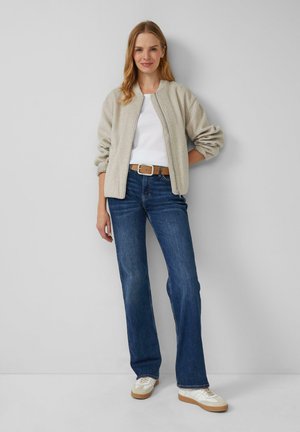 Veste bomber zippée gris clair sur un t-shirt blanc, associée à un jean évasé bleu et à des baskets blanches avec des semelles beige. Look simple et décontracté.