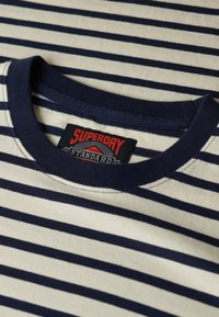 Navy- und creamfarbener gestreifter Baumwoll-T-Shirt mit geripptem Rundhalsausschnitt und einem roten Markenzeichen, das nahe dem Kragen eingenäht ist.