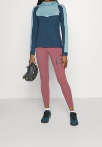 Långärmad marinblå och ljusblå atletisk topp med hög krage, tillsammans med rosa leggings som har en mörk logotyp, och svarta klätterskor.