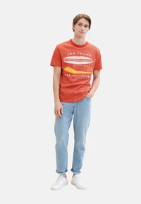 Camiseta a rayas rojas con un estampado gráfico, combinada con jeans azul claro y zapatillas blancas. La camiseta tiene un cuello redondo y mangas cortas.