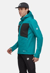 Ein Mann trägt eine türkis-schwarze Kapuzenjacke und schwarze Hosen mit Logos, steht vor einem schlichten weißen Hintergrund und blickt nach links.