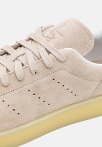 Zapatilla de ante beige con cordones planos, detalles perforados y suela de goma amarilla. Presenta un sutil logo grabado en el talón.