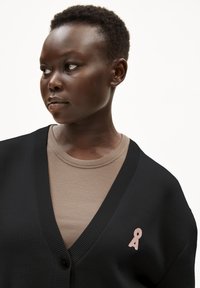 Svart kofta med djup V-ringning och ribbad textur, prydd med en rosa broderad detalj. Lager på en beige ribbad topp.