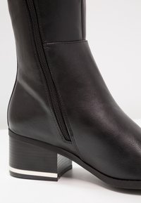 Bottine en cuir noir avec une fermeture éclair sur le côté, bout carré et un petit talon bloc avec une rayure d'accent blanc. Texture lisse sur toute la surface.