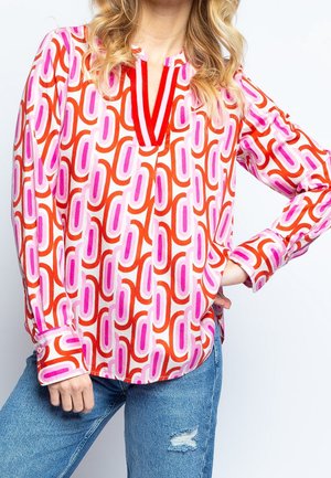 Mujer que lleva una blusa de manga larga con un patrón abstracto de cadenas en rojo, rosa y naranja y vaqueros azules con un pequeño desgarro en la rodilla izquierda.