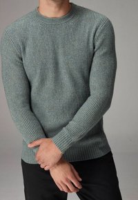 Helleblaue, gerippte Strickjacke mit langen Ärmeln, rundem Halsausschnitt und strukturiertem Stoff. Die Hände sind an der Taille gekreuzt, kombiniert mit schwarzen Hosen.