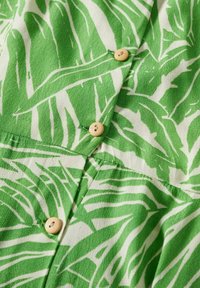 Tissu vert et blanc avec un motif de feuilles tropicales et trois boutons brun clair cousus en rangée verticale sur le vêtement.