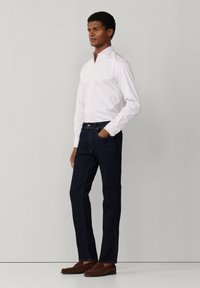 Muž stojaci s jednou rukou v vrecku, oblečený v bielej košeli na gombíky, tmavomodrých džínsoch a hnedých mokasínach na jednofarebnom pozadí.