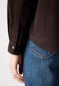 Person wearing dark brown long-sleeve shirt and blue denim jeans with visible back pocket, hand relaxed at side against light background.