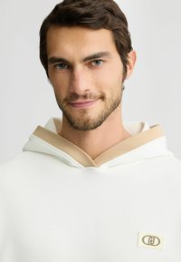 Weißer Hoodie mit beigem Kordelzug und Besatz, mit einem Logo-Patch auf der Brust. Glatte Textur und lockerer Schnitt.