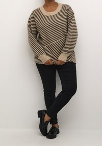 Pull beige et marron à rayures avec larges poignets, associé à un pantalon skinny noir et des mocassins noirs brillants, posé devant un fond blanc.