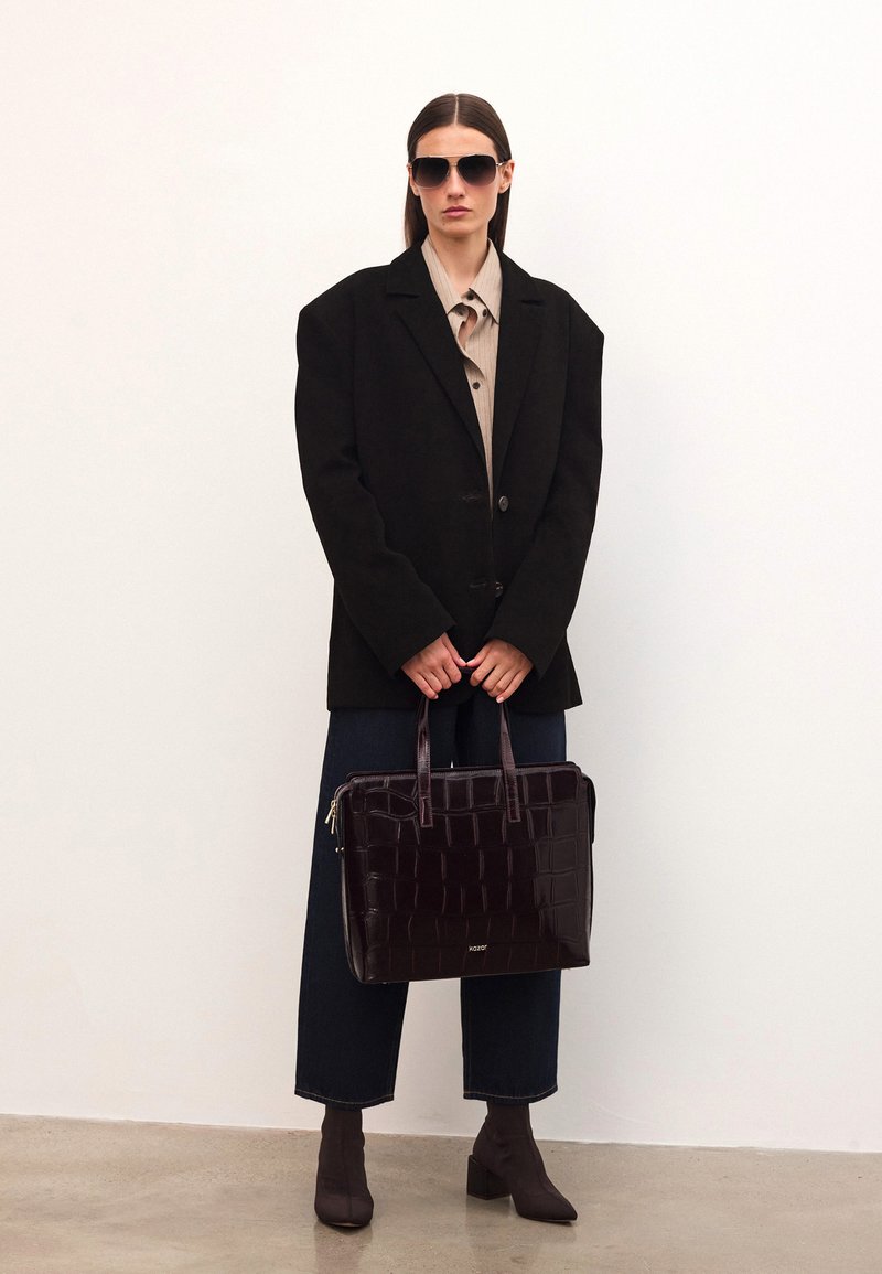 Tumma ruskea nahkakäsilaukku, jossa on teksturoitu krokotiilikuosi, suorakulmainen muoto, yläkahvat ja vetoketjukiinnitys. Mallilla on päällään suuri musta bleiseri.