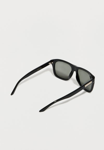Μαύρα γυαλιά ηλίου με ορθογώνιο πλαίσιο και σκούρους φακούς, που ακουμπούν σε λευκή επιφάνεια, φαίνονται από πίσω δεξιά πλευρά.