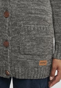 Grauer Strickcardigan mit strukturiertem Muster, Holzknöpfen, einer Vordertasche und einem gebrandeten Lederpatch am Saum.