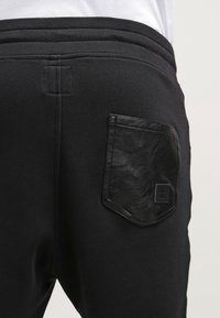 Svarta mjukisbyxor med en texturerad svart läderficka bak. Tyget är mjukt, med ett brett elastiskt midjeband och sydda sömmar.