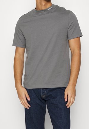 Mand iført en ensfarvet grå kortærmet t-shirt med Calvin Klein-logo på kraven og mørkeblå jeans, stående mod en hvid baggrund.