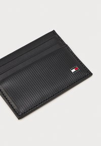 Μαύρος δερμάτινος θήκη καρτών με ριγέ υφή, τέσσερις θέσεις για κάρτες και μικρή λογότυπη λεπτομέρεια σε κόκκινο, άσπρο και μπλε.