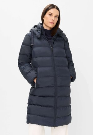 Frau trägt einen langen, marineblauen, gesteppte Daunenmantel mit Kapuze und steht mit den Händen in den Taschen vor einem schlichten, hellen Hintergrund.