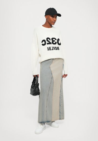 White oversized sweater with black text, beige and blue patchwork denim skirt, white sneakers, and a black cap. Textured handbag with accents.