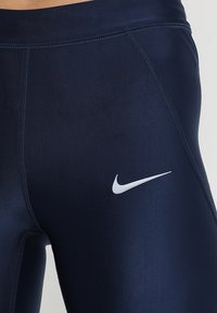 Leggings atletici blu navy realizzati in tessuto elasticizzato, con un logo bianco di Nike sul lato sinistro e dettagli con cuciture piatte.