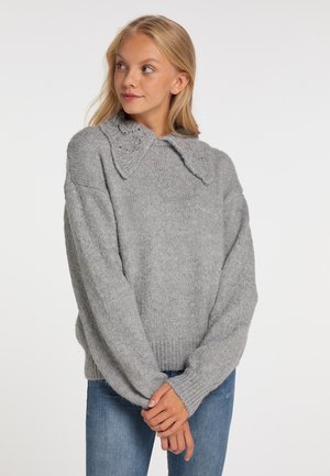 Ung kvinde med blondt hår iført en lysegrå striktrøje med en bred krave og blå jeans, stående med foldede hænder.