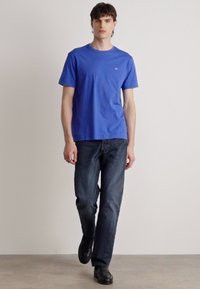 Blaue Baumwoll-T-Shirt kombiniert mit dunklen Jeans. Das Shirt hat einen Rundhalsausschnitt und kurze Ärmel; dezentes Logodesign auf der Brust.