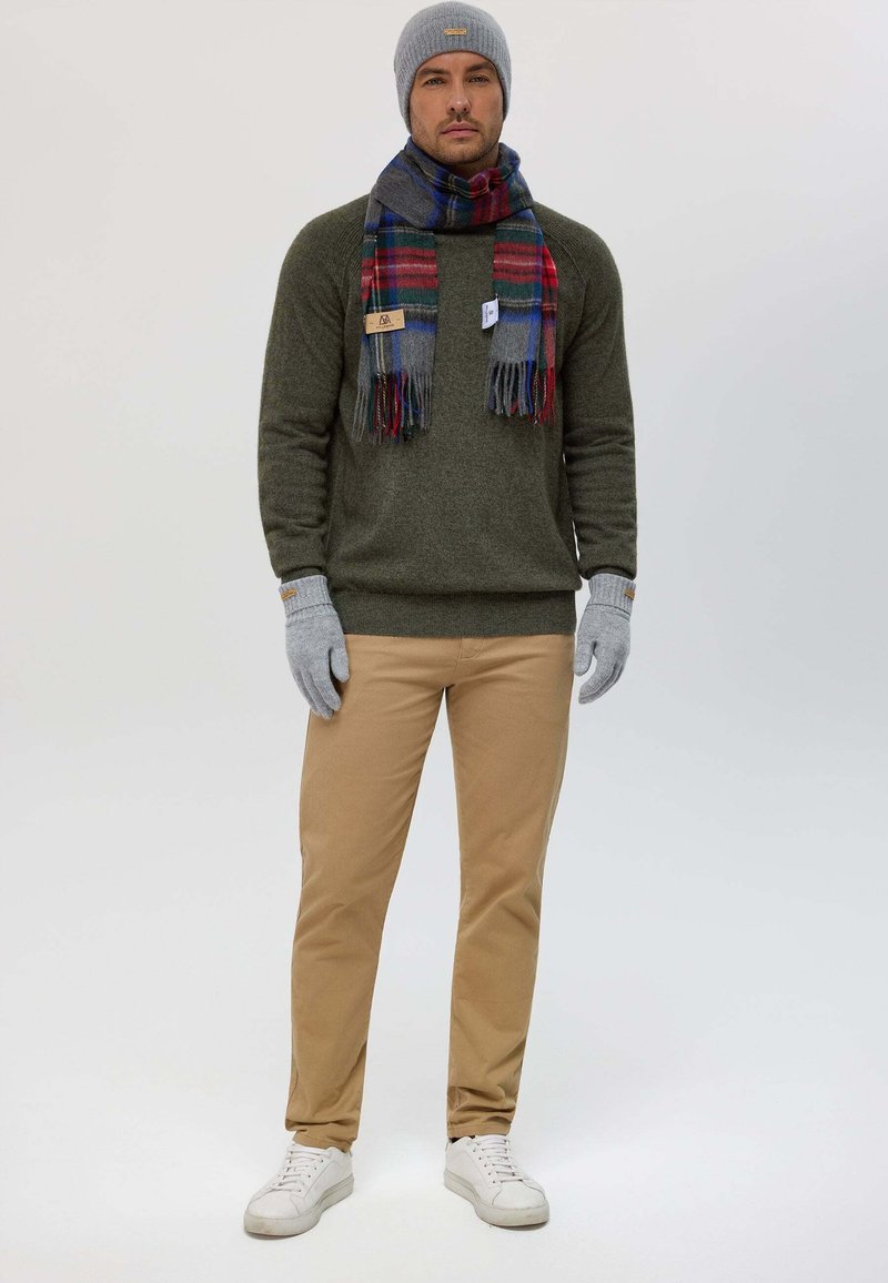 Bonnet gris et écharpe à carreaux bleus et rouges, pull vert, gants gris, pantalon beige et baskets blanches. Tenue d'hiver décontractée.