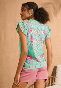 Green floral top with ruffled sleeves, featuring pink and orange patterns. Paired with light pink shorts, showcasing a relaxed fit.