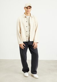 Svetlo béžová bunda na gombíky cez béžovú košeľu, skombinovaná s tmavými šedými nohavicami a bielymi teniskami. Tento look je neformálny a uvoľnený.