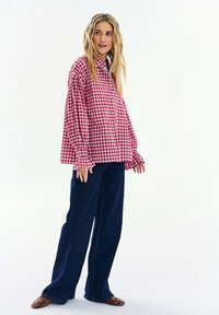 Žena s dlouhými blond vlasy, která má na sobě červenobíle kostkovanou volnou košili, tmavé široké kalhoty a boty se vzorem leopardí kůže, stojící před bílým pozadím.