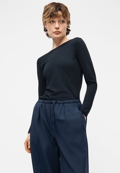 Νεαρό άτομο με κοντά μαλλιά, φοράει εφαρμοστό μαύρο μακρυμάνικο τοπ και φαρδιά σκούρα μπλε παντελόνια με τα χέρια στις τσέπες, κοιτάζοντας προς τα αριστερά.