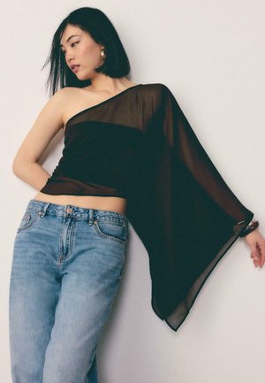 Młoda kobieta o krótkich czarnych włosach nosi czarny, przezroczysty top z odsłoniętym jednym ramieniem i niebieskie dżinsy, pozując na jasnym, jednolitym tle.