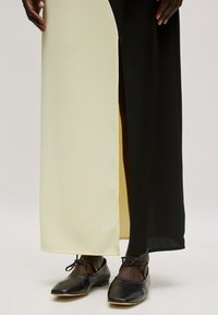 Maxi-jurk met een split ontwerp en zwarte en pastelgele panelen, gemaakt van een zachte stof. Zwarte schoenen met een strikdetail worden gedragen.