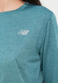 Τζελ αθλητική μπλούζα από υφασμάτινη υφή, με στρογγυλή λαιμόκοψη και λευκό λογότυπο New Balance στο αριστερό μέρος του στήθους.