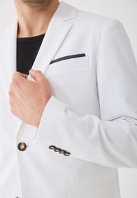 Biała marynarka z klapami, czarnym detalem na kieszeni i czterema guzikami na mankietach. Gładka faktura i dopasowany krój.