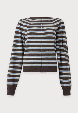 Femme portant un pull rayé noir, blanc et bleu avec un jean bleu clair à jambes larges et des chaussures marron, debout avec une main dans la poche.