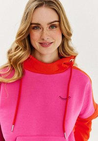 Mikina s kapucí růžového těla a oranžovými rukávy, s dnes oblékací šňůrkou a malým černým designem řas na přední části.