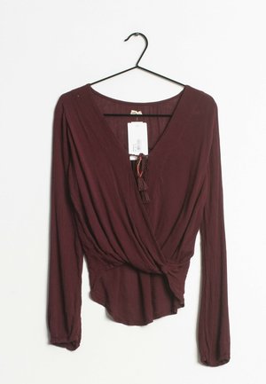 Blusa bordeaux a maniche lunghe con scollatura a V e dettaglio frontale annodato. Realizzata in tessuto testurizzato, presenta un'etichetta con informazioni su prezzo e marca.
