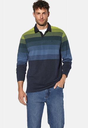 Mann in einem langärmligen, kragenlosen Hemd mit horizontalen grünen und blauen Streifen, kombiniert mit blauen Jeans, steht mit einer Hand in der Tasche.
