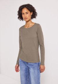 Kvinna med lockigt mörkt hår, iklädd en taupe långärmad skjorta och ljusblå jeans, står mot en platt vit bakgrund.