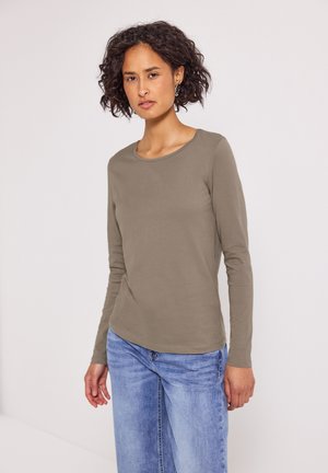 Frau mit lockigem, dunklem Haar, die ein taubfarbenes Langarmshirt und hellblaue Jeans trägt, steht vor einem schlichten weißen Hintergrund.