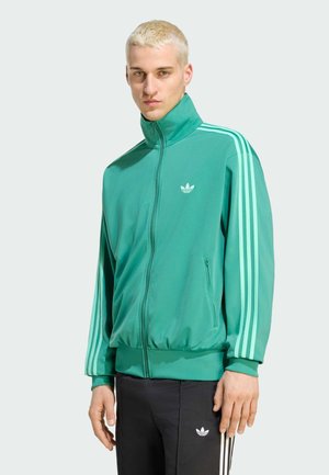 Jeune homme aux cheveux blonds courts portant une veste de survêtement verte Adidas à bandes blanches et un pantalon de survêtement noir Adidas.