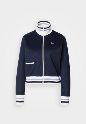 Navy zip-up takki, polyesteriseos, korkea kaulus raidallisella resorilla, valkoiset yksityiskohdat, etupussit, Lacoste-logo rinnassa.